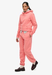 Sudadera rosa con un bolsillo frontal, cordones y puños de canalé, combinada con joggers a juego y mocasines oscuros. Tela texturizada visible.
