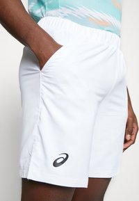 Fehér sportnadrág könnyű anyagból, rugalmas derékkal, oldzsebekkel és fekete logó díszítéssel az alján.