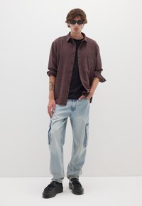 Camicia a scacchi bordeaux sopra una maglietta nera, abbinata a jeans cargo azzurri chiari e scarpe nere. I capelli sono ondulati e sono indossati occhiali da sole.