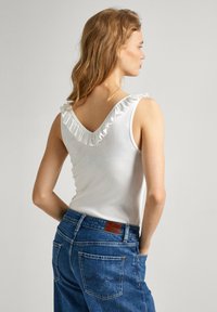 Top bianco senza maniche con bordo arricciato attorno a un profondo scollo a V, abbinato a jeans in denim blu con tasche posteriori e una patch in pelle marrone.