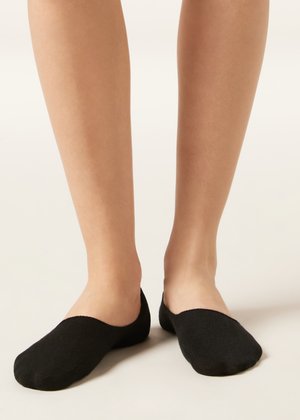 Piernas con calcetines negros de corte bajo y tipo invisibles, sobre un fondo claro y neutro.