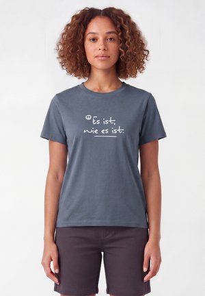 Vrouw met krullend haar die een blauwgrijs T-shirt draagt met Duitse tekst "Es ist, wie es ist." en donkere korte broek tegen een effen lichte achtergrond.