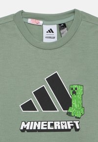 Zeleni bombažna majica s kratkimi rokavi, ki prikazuje grafiko pikseliziranega Creeperja iz igre Minecraft, pod njim pa kontrastno črno Adidas logo in ime blagovne znamke.