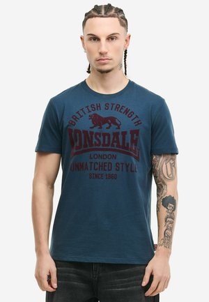 Mladý muž s vrkočmi, oblečený v modrom tričku Lonsdale s hnedým nápisom a logom leva, ukazuje tetovania na pravom ramene.