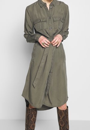 Personne portant une robe boutonnée vert olive avec poches poitrine et ceinture nouée, associée à des bottes montantes en cuir de serpent et des bagues dorées aux doigts.
