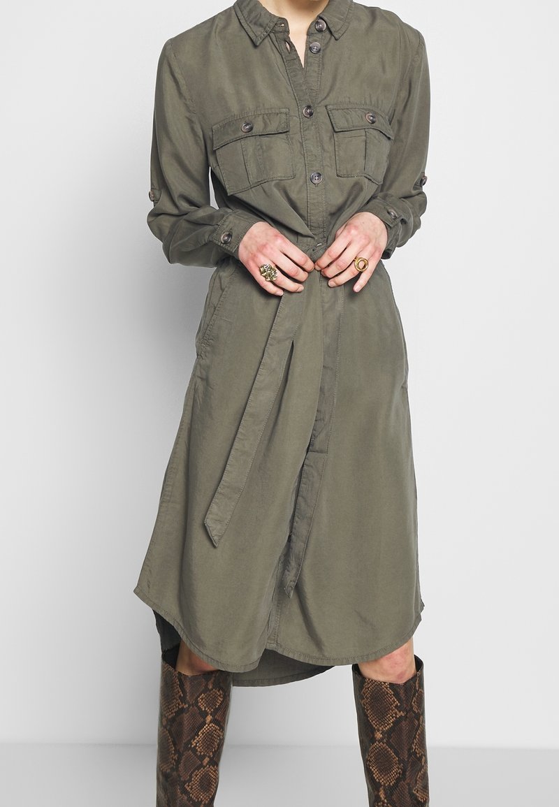 Personne portant une robe boutonnée vert olive avec poches poitrine et ceinture nouée, associée à des bottes montantes en cuir de serpent et des bagues dorées aux doigts.