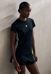 Musta urheilupaita lyhyillä hihoilla, jossa valkoinen Adidas-logo ja raidat sivuilla. Yhdistetty samansävyisten mustien shortsien kanssa. Suloinen yksityiskohta.
