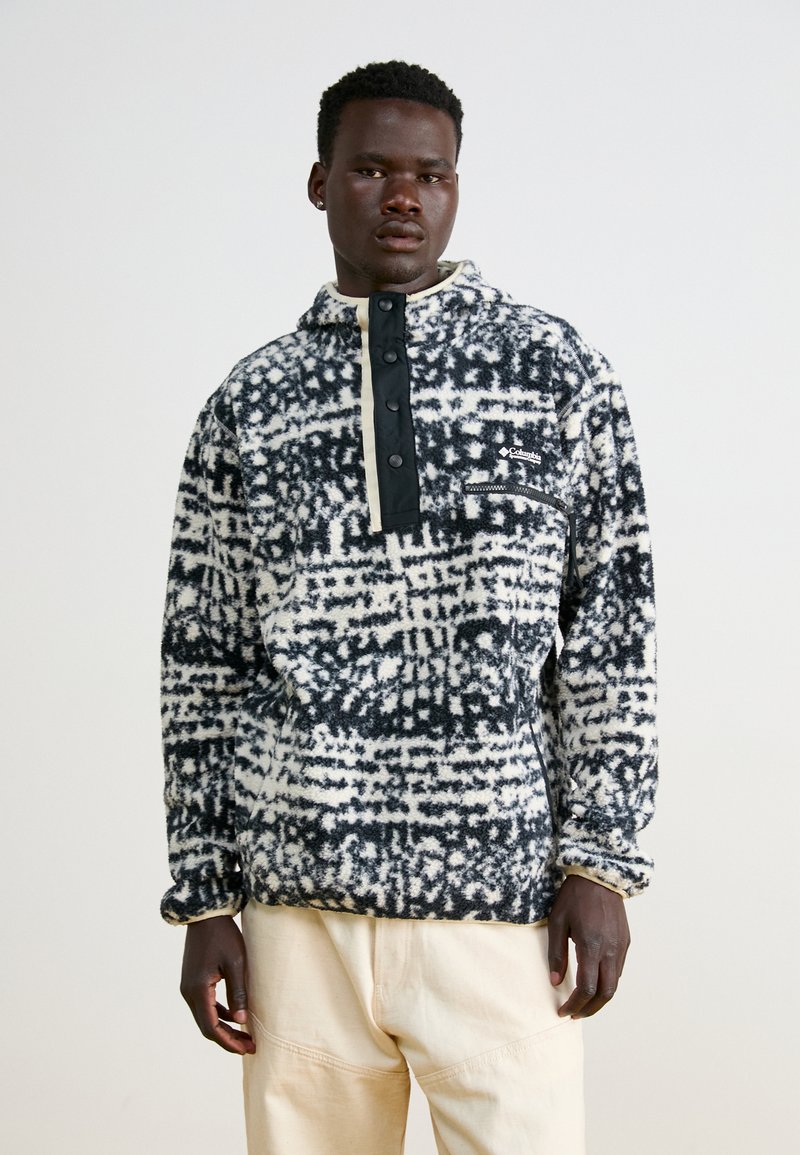 Polarowy pulower w czarno-białym wzorze, z półzamkiem z przodu, kapturem i kieszenią na piersi. Miękka tekstura, luźny krój.