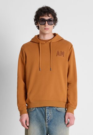 Giovane uomo con felpa marrone con cappuccio con "AM" sul petto e jeans blu scoloriti, in piedi davanti a uno sfondo chiaro uniforme.