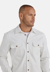 Weißes Button-up-Hemd mit zwei Brusttaschen, braunen Knöpfen und einem Kragen. Der Stoff wirkt glatt mit einer leichten Textur.