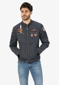 Dark blue bomber jacket with zip front, stand collar, and multiple embroidered patches including a flag and brand logos. Features side pockets.