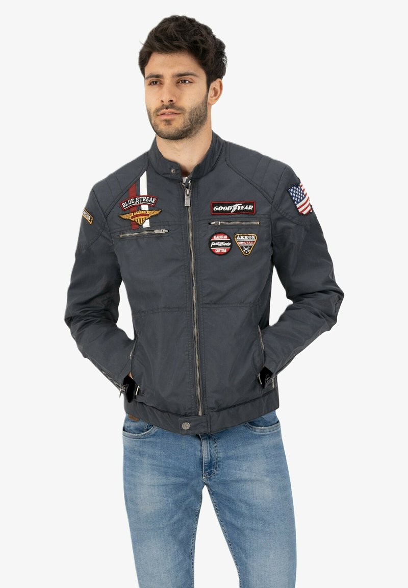 Dark blue bomber jacket with zip front, stand collar, and multiple embroidered patches including a flag and brand logos. Features side pockets.