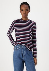 Langermet stripete skjorte i marineblå, hvit og rød, med rund hals og en liten grønn logo. Kombinert med høyt midjebånd blå jeans.