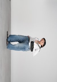 Femeie îmbrăcată cu o cămașă albă cu nasturi, blugi evazați albaștri, sandale negre și o geantă neagră pe umăr, pe un fundal gri simplu.