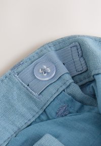 Tissu bleu clair légèrement texturé, avec un bouton en plastique transparent et des accents cousus, présentant une zone de col pliée.