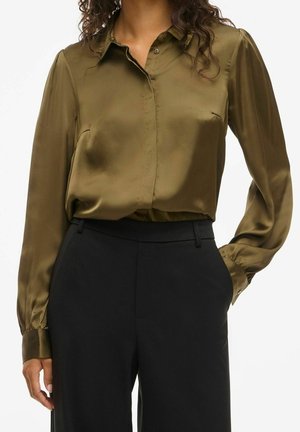 Frau trägt eine glänzende, olivgrüne Bluse mit langen Ärmeln, die in hoch taillierte schwarze Hosen gesteckt ist, mit einer Hand in der Hosentasche.