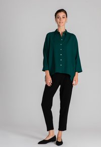 Blusa verde con bottoni, dalla vestibilità ampia, maniche lunghe e orlo curvo; abbinata a pantaloni neri a sigaretta e scarpe nere slip-on.