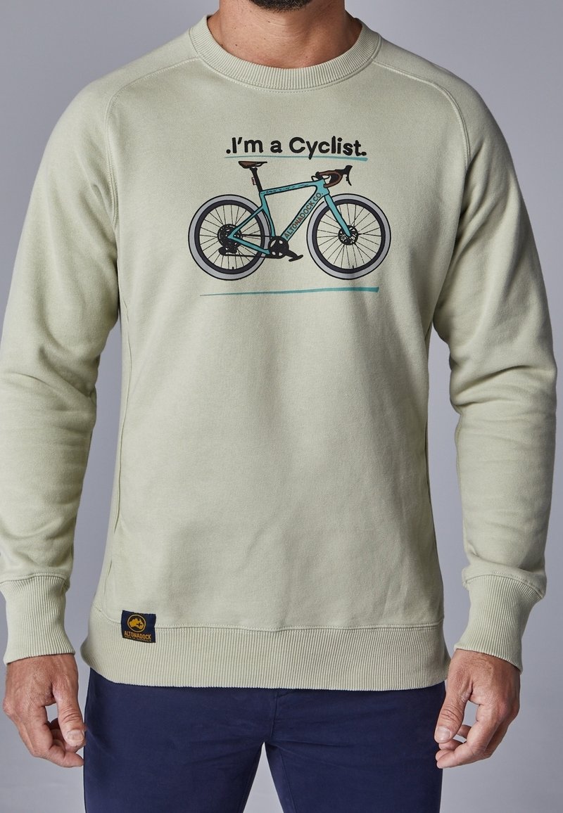 Sweatshirt beige avec le texte « Je suis cycliste » et un motif de vélo turquoise, porté par une personne portant un pantalon marine devant un fond gris uni.