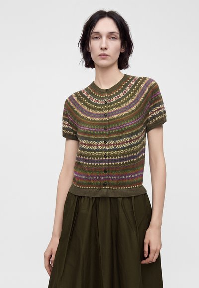 Femme portant un cardigan en maille à manches courtes boutonné avec des motifs géométriques multicolores et une jupe vert olive foncé.