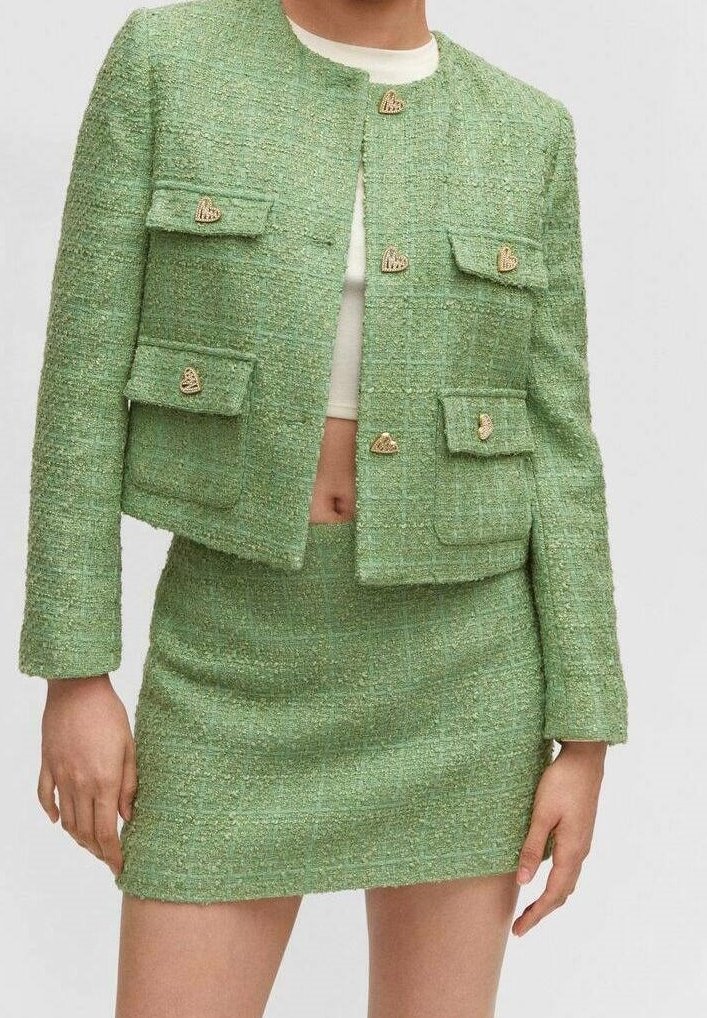 Femme portant une veste courte verte texturée avec des boutons dorés en forme de cœur et une mini-jupe taille haute assortie sur un crop top blanc.