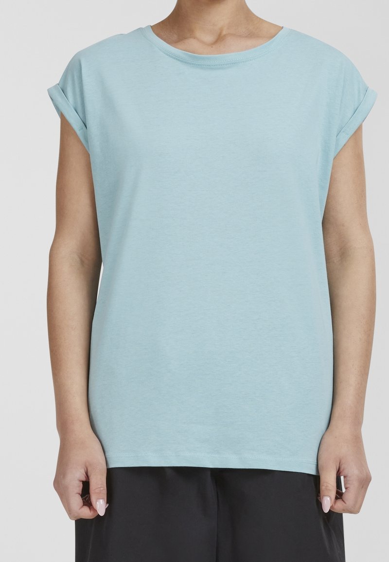 Personne portant une chemise bleu clair à manches courtes, avec les poignets retroussés, et un pantalon noir sur un fond uni clair.