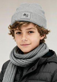 Gray ribbed beanie with a rectangular patch, paired with a thick, braided gray scarf, worn over a black puffer jacket. Subtle texture details visible.