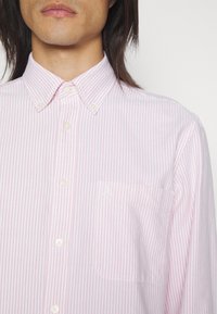 Chemise à rayures rose et blanches avec boutons, dotée d'un col, d'une poche avant et d'une broderie de logo discrète. Le tissu semble lisse et léger.