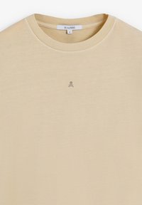 Camiseta de cuello redondo beige hecha de un tejido suave, con un pequeño emblema de calavera oscura en el pecho. Sin patrones ni acentos adicionales visibles.