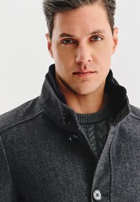 Ciemnoszary płaszcz w jodełkę z wysokim kołnierzem, ozdobiony dużymi czarnymi guzikami. Pod spodem widać teksturowany zielony sweter z dzianiny.