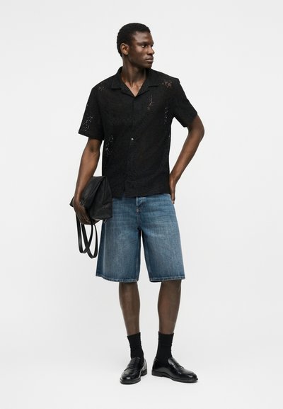 Άνδρας με μαύρο δαντελένιο κοντομάνικο πουκάμισο, σορτς από τζιν μέχρι το γόνατο, μαύρες κάλτσες και μοκασίνια, κρατώντας μια μαύρη τσάντα σε ένα χέρι.