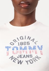 Ribana krem T-shirt s višebojnim grafičkim printom s natpisom "TOMMY" u plavoj i crvenoj boji, te "ORIGINAL 1985 JEANS NEW YORK" u crnoj boji.