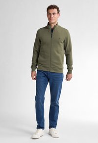 Olijfgroene zip-up sweatshirt met een textuurpatroon, elastische manchetten en zoom, gecombineerd met blauwe jeans en witte sneakers.