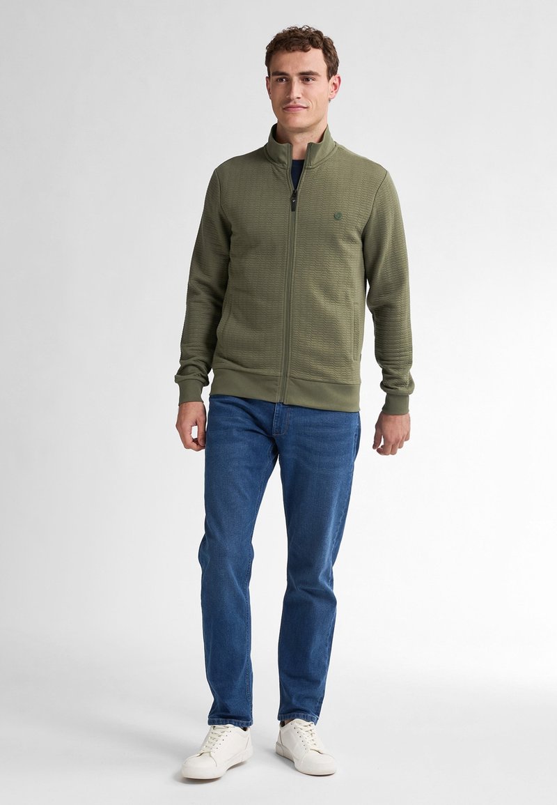 Oliwkowy sweatshirt zapinany na zamek z teksturowanym wzorem, z prążkowanymi mankietami i dołem, w połączeniu z niebieskimi dżinsami i białymi sneakersami.