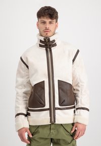 Bílá bunda z ovčího rouna s hnědými koženými akcenty, vysokým límcem a dvěma velkými předními kapsami. Obsahuje zipové zapínání a kontrastní detaily.
