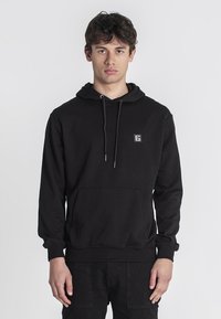 Sudadera negra de material suave, con un bolsillo frontal, capucha con cordones y un pequeño parche con el logo en el pecho.
