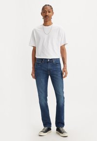 Muž s opletenými vlasmi, oblečený v bielom tričku, modrých džínsoch, čiernych teniskách a zlatom náhrdelníku, ktorý stojí pred plain bielym pozadím.