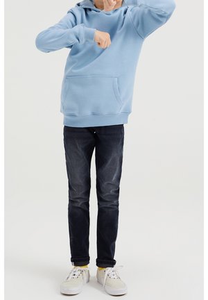 Sudadera con capucha de color azul claro, con un bolsillo frontal, combinada con jeans de mezclilla oscura y zapatillas blancas con detalles amarillos. Ajuste y diseño casual.