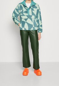 Sudadera de forro polar en tonos azul verdoso y menta con patrones geométricos, combinada con pantalones verdes de cuero sintético y zapatos de plataforma de color naranja brillante.