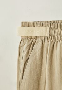 Pantaloni din țesătură beige, având un brâu texturat cu o bandă elastică de culoare deschisă. Designul plissetat sporește volumul și căderea.