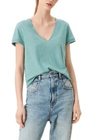 Helles Teal T-Shirt mit V-Ausschnitt und kurzen umgeschlagenen Ärmeln, gefertigt aus einem weichen, atmungsaktiven Material. Kombiniert mit hoch geschnittenen hellblauen Jeans.