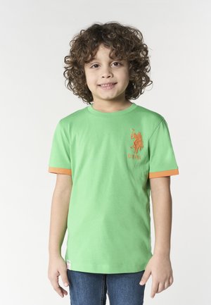 Ragazzo giovane con capelli ricci che indossa una maglietta verde con bordi arancioni e logo USPA, in piedi davanti a uno sfondo chiaro semplice.