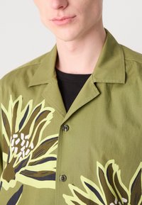Zöld, rövid ujjú ing, nagy virágmintával sárgában, barnában és fehérben. Galléros nyakkivágással és fekete gombos záródással rendelkezik.