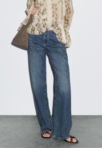 Leichte, gemusterte Bluse in Creme mit Rüschen, kombiniert mit dunkelblauen, weiten Jeans. Ergänzt mit einer beigen Tasche und flachen Sandalen.