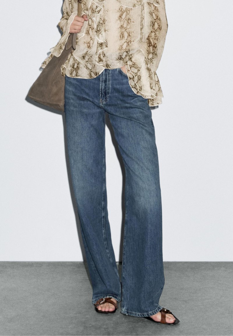 Leichte, gemusterte Bluse in Creme mit Rüschen, kombiniert mit dunkelblauen, weiten Jeans. Ergänzt mit einer beigen Tasche und flachen Sandalen.