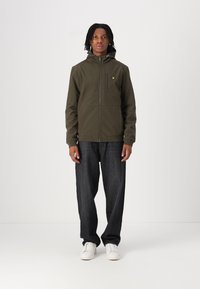 Oliven grønn hettejakke med glidelås, med et logo på brystet, paired med mørke jeans og hvite joggesko. Minimalistisk design og glatt tekstur.
