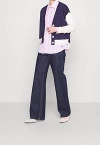 Marinblå och krämfärgad färgblockerad kofta över en rosa och vit randig skjorta. Mörk denimbyxor med vida ben och strukturerade, ljusrosa skor.