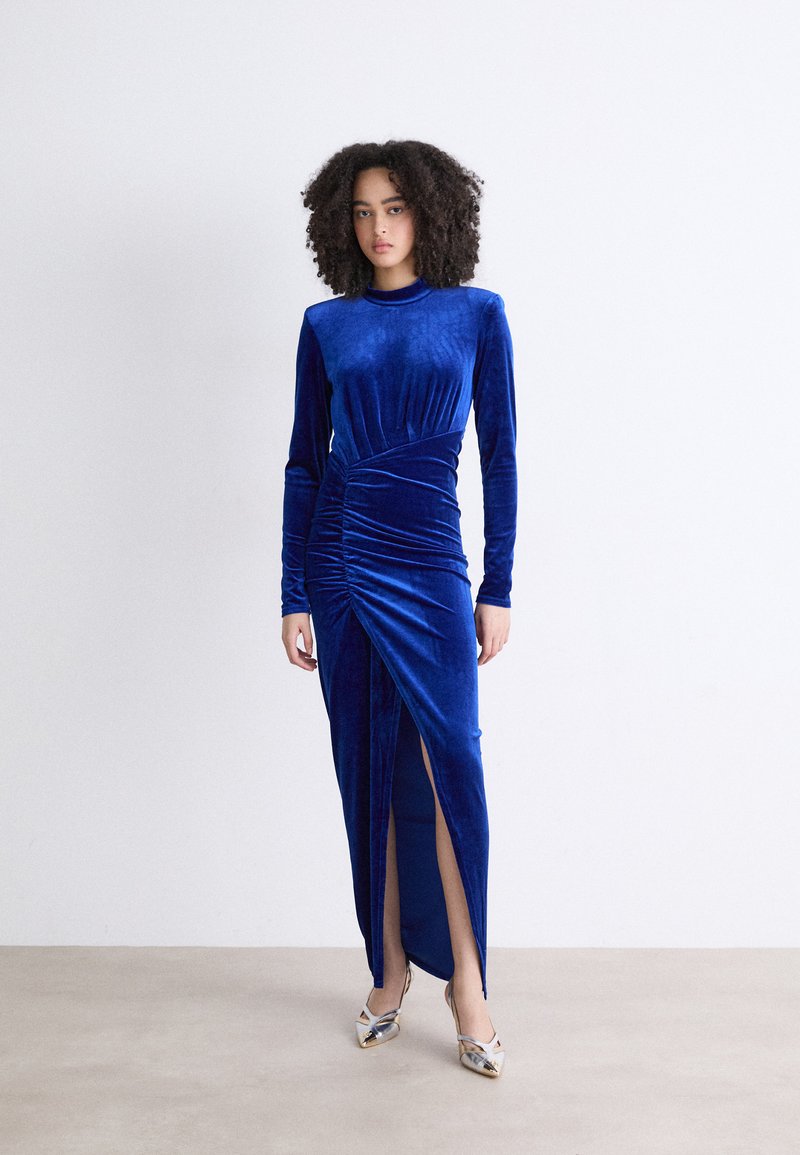 Vestido de veludo azul de mangas compridas com detalhes franzidos e uma fenda alta, combinado com sapatos de salto alto metálicos. Textura suave e luxuosa.