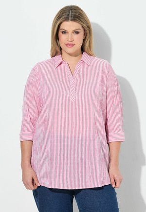 Kobieta o blond włosach, ubrana w różowo-białą koszulę w pionowe paski z rękawami do łokcia oraz niebieskie dżinsy, stojąca na białym tle.