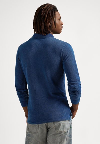 Μπλε ναυτικών μακρυμάνικη μπλούζα πόλο με υφασμένο πλέξιμο, κλασικό γιακά και πλευρικές σχισμές, συνδυασμένη με ανοιχτό γκρι τζιν.