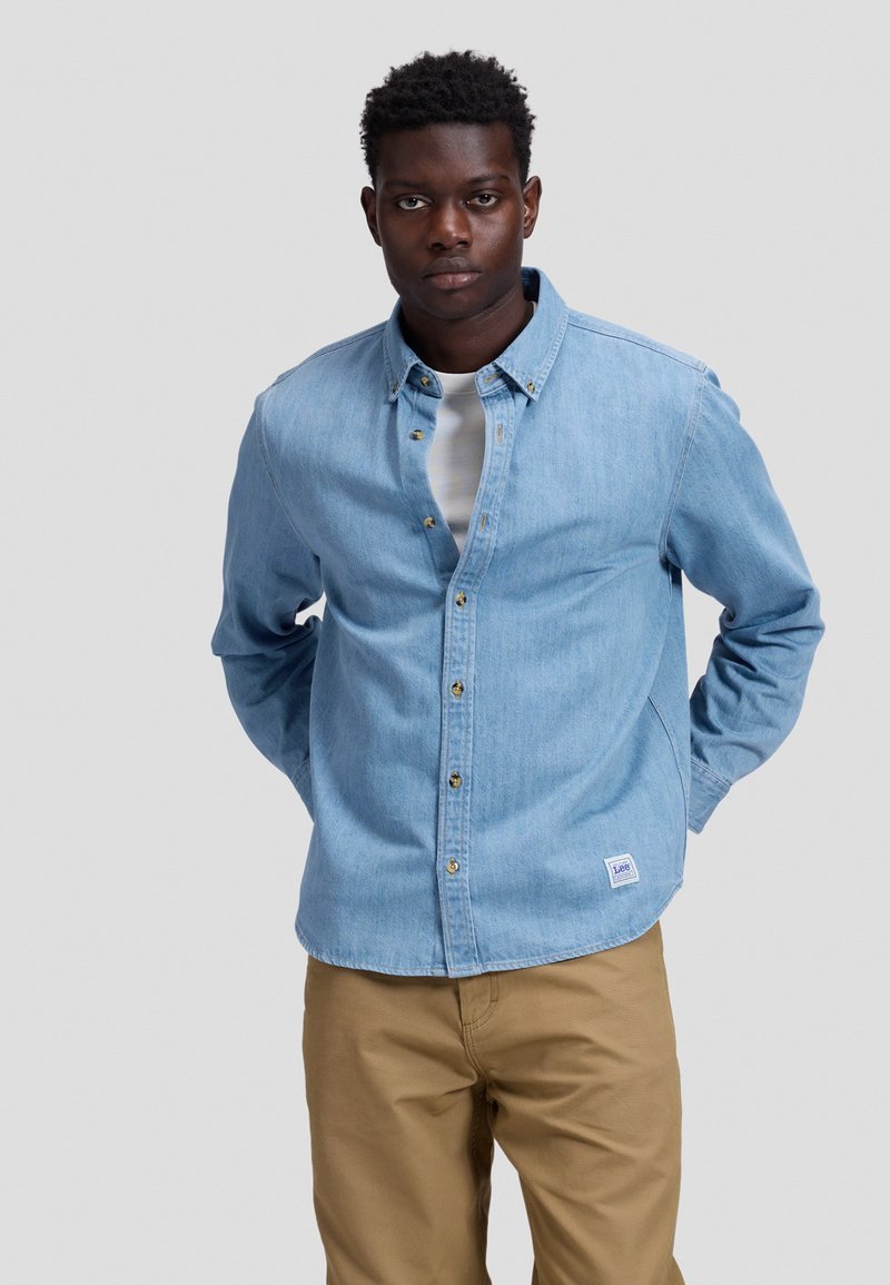 Jeune homme portant une chemise en jean bleu clair boutonnée par-dessus un t-shirt blanc et un pantalon beige, debout, les mains dans le dos.